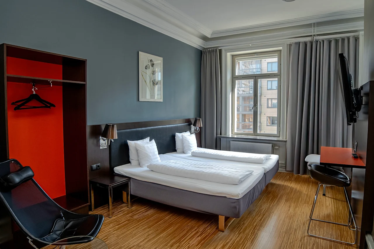 Ett modernt hotellrum med två sängar, ett fönster med grå gardiner, en garderob och en liten skrivbordsplats.