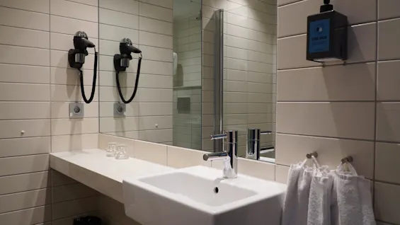 A white bathroom sink with a chrome faucet sits against a tiled wall. Above, a mirror reflects a hairdryer mounted beside it. A "HAND WASH" dispenser is on the wall. Towels hang nearby.