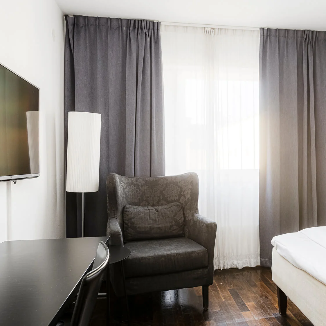 En mörkgrå fåtölj vätter mot ett fönster täckt av tunna och ogenomskinliga gardiner i ett minimalistiskt sovrum, kompletterat med en säng och en väggmonterad TV.