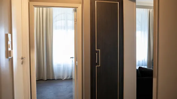 En hotelgang med en åben dør, der afslører et lyst værelse, og en lukket mørk trædør med elegante gulddetaljer.