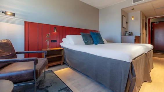 A modern hotel room with a large bed, red wall panels, and a brown leather armchair.