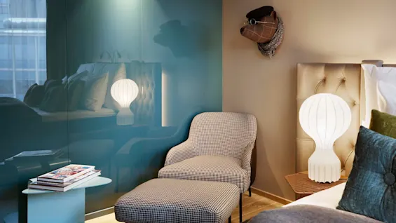 A checkered armchair with ottoman is placed beside a bed in a cozy room. A modern lamp sits on a side table, with decorative animal head on the wall.