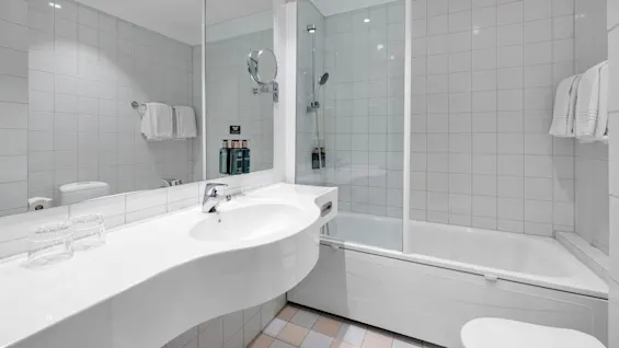 Et hvidt badeværelse med en buet vaskebænk, glasdør til brusen, vægmonteret spejl og håndkleholder. Flasker mærket "Body Lotion," "Shampoo," og "Conditioner" står ved vandhane.