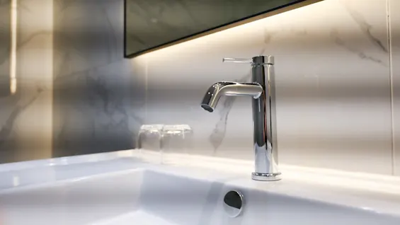 En moderne baderomsvask med en elegant kromkran, to glass og marmormønstrede fliser med bakgrunnsbelysning.