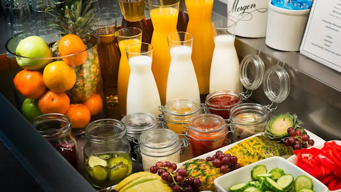 A variety of fresh fruits, milk in glass carafes, and condiments in jars displayed for a buffet breakfast in a well-lit setting.