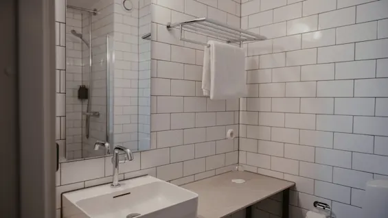 Et rent, moderne badeværelse med hvide fliser, en elegant håndvask og en bruser synlig i spejlet.