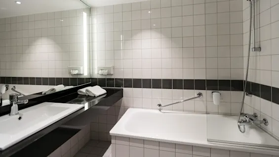 A modern bathroom features a white tiled bathtub with a shower, flanked by a large mirror and sink on the left. Towels are neatly placed on the counter.