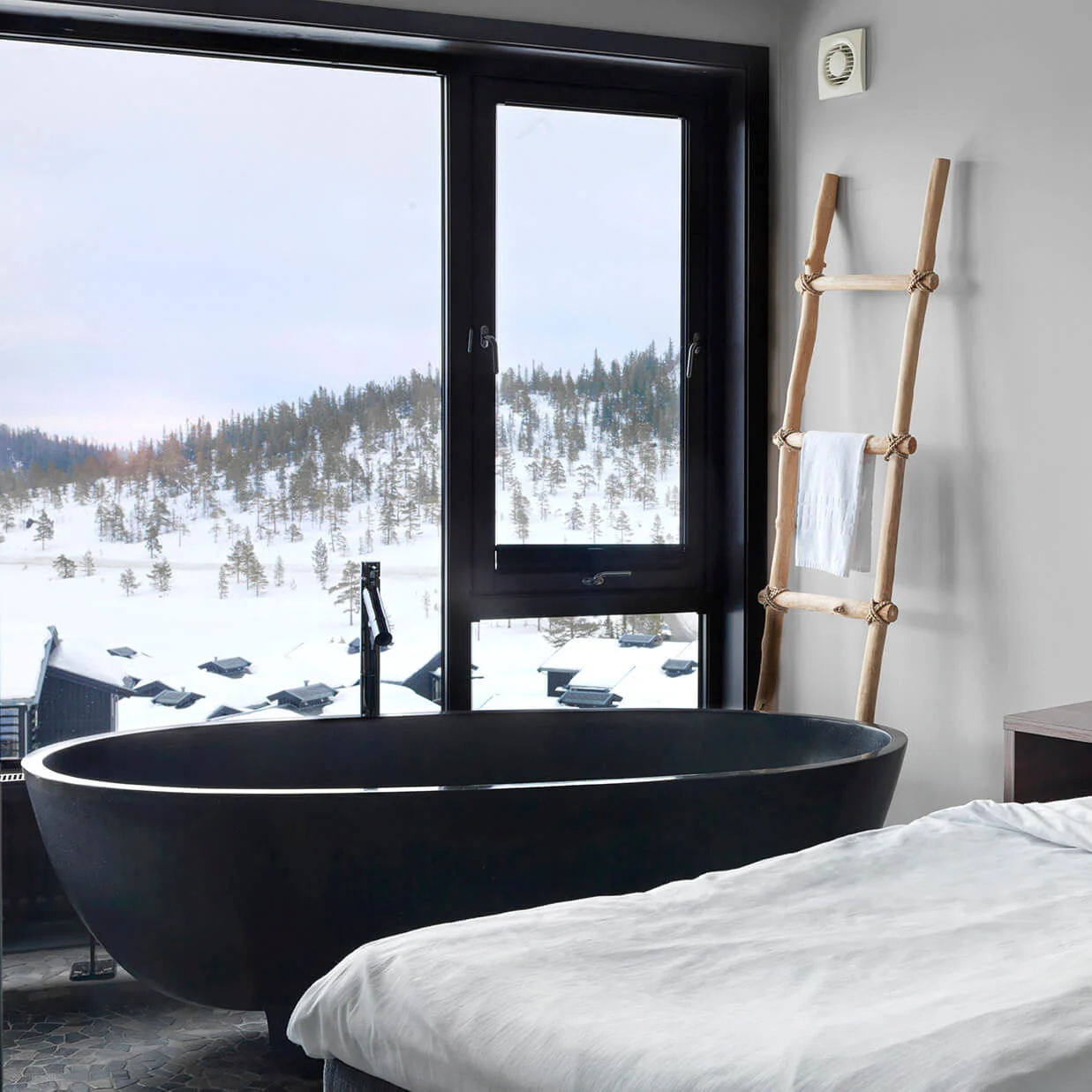 A modern bedroom features a bed, a freestanding bathtub, and a wood ladder with a towel, set against a window showcasing a snowy landscape and forested hills.