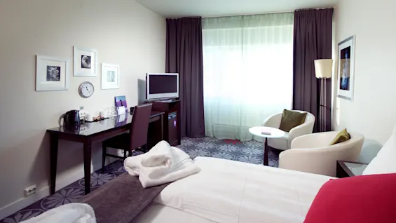 A modern hotel room with a neatly made bed, work desk, television, and seating area, illuminated by natural light through sheer curtains.