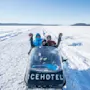Snescooter mærket "ICEHOTEL" med tre personer vinkende, kørende gennem et snedækket landskab under klare blå himmel.