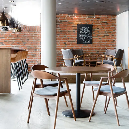 En hyggelig café har træborde med stole omkring. Bardisk med høje stole til venstre; hylder og dekorationer på væggene. Murstensvæg tilføjer varme.
