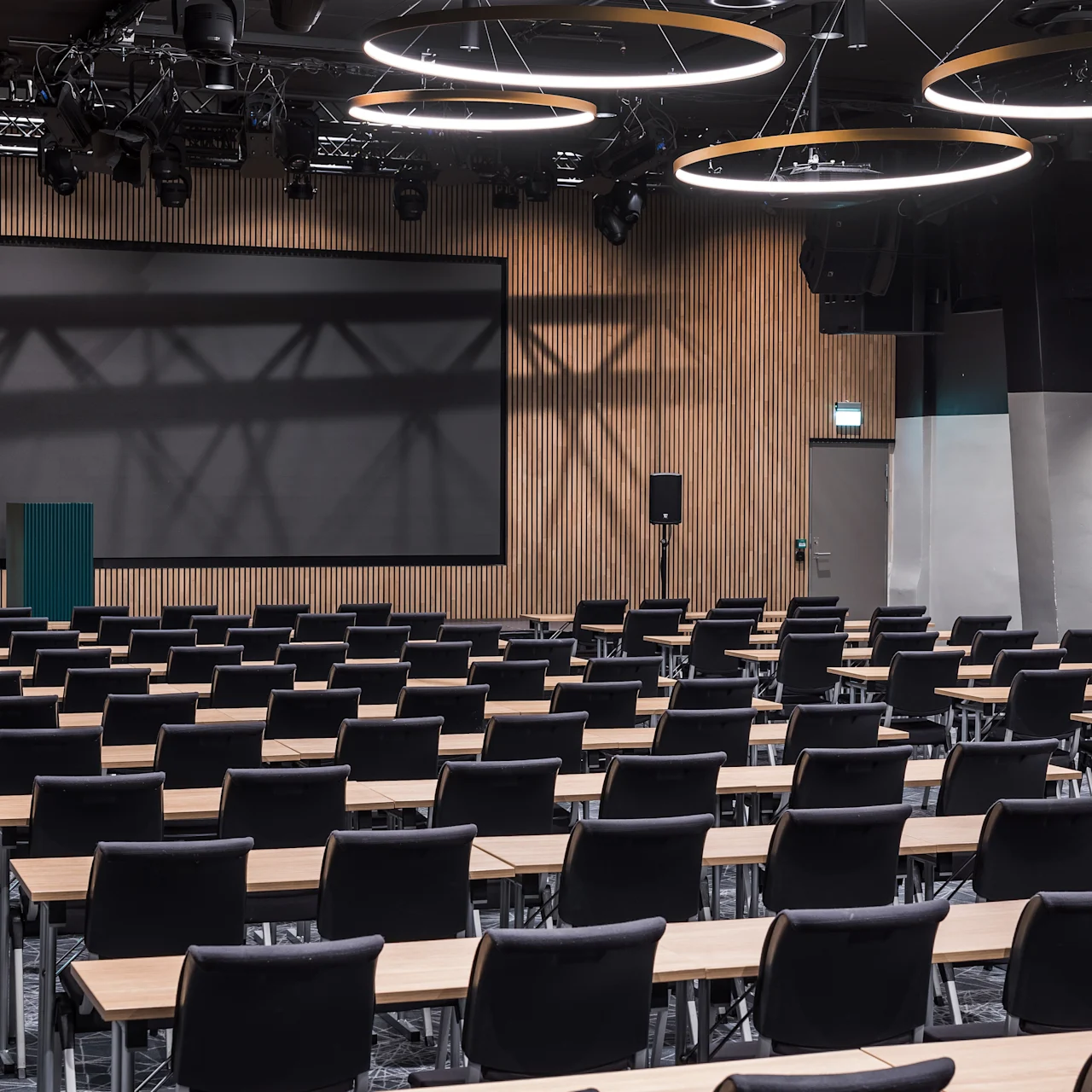 Konferenssihuone, jossa on riveittäin pöytiä ja tuoleja, jotka ovat suunnattu kohti suurta näyttöä puupaneeliseinällä. Pyöreät valot roikkuvat katosta valaisten huonetta.