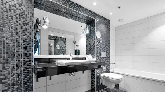 Vaskens spejlbillede viser elegant badeværelse; vægmonteret toilet og oplyst vask. Stilige sorte og hvide fliser dominerer. Væghylde med toiletartikler. Badekar i baggrunden fuldender et moderne indtryk.