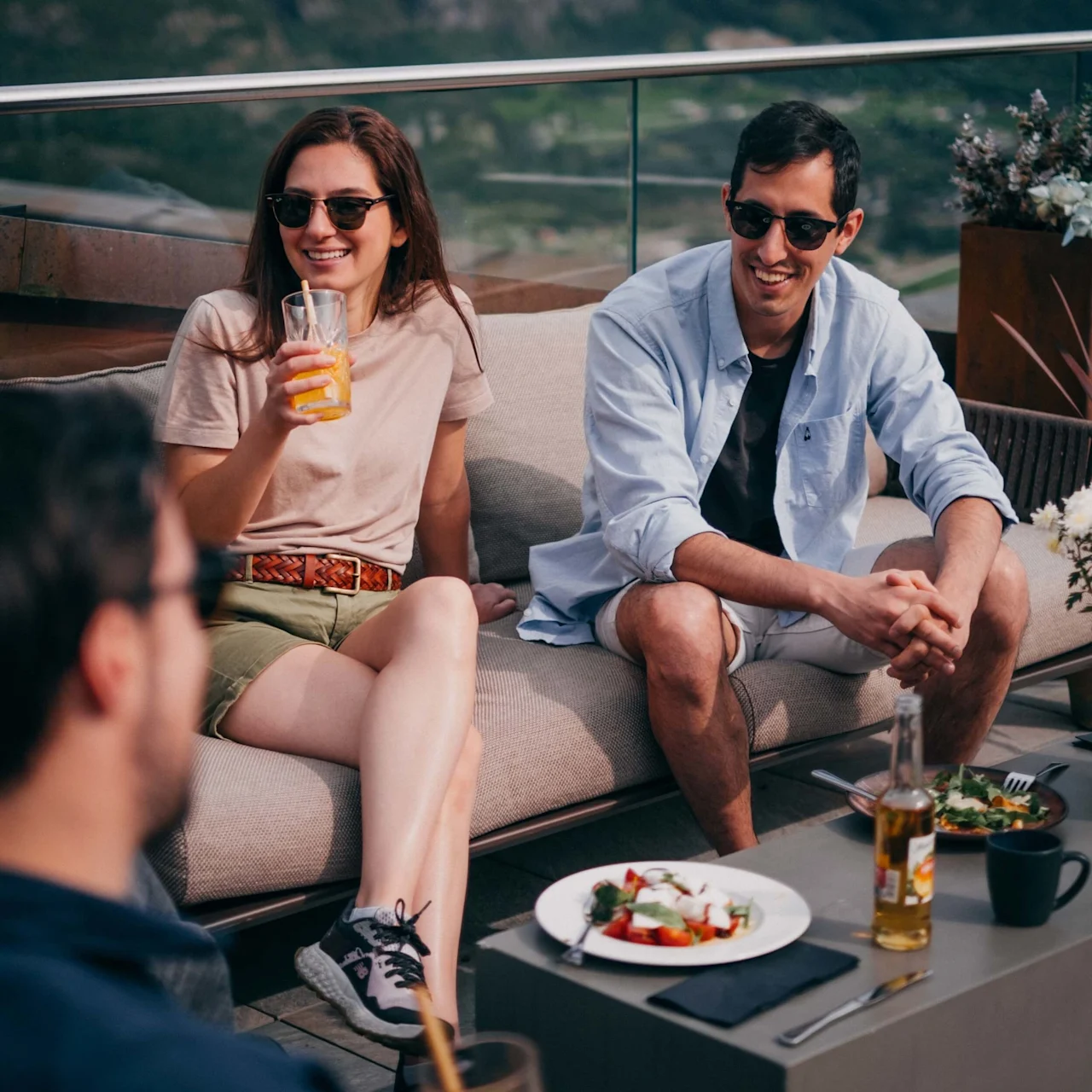 A woman holding a drink and a man smiling sit on an outdoor couch conversing with another person; a table with food and drinks is in front of them.
