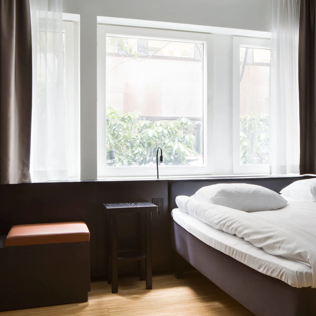 Seng med hvide lagener er pænt reddet, placeret ved siden af store vinduer med hvide gardiner, sollys strømmer ind i det moderne, minimalistiske rum.