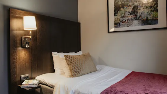 A welcoming single hotel room featuring a bed with a purple throw, a reading lamp, and a framed artwork. Home Hotel Kung Oscar.