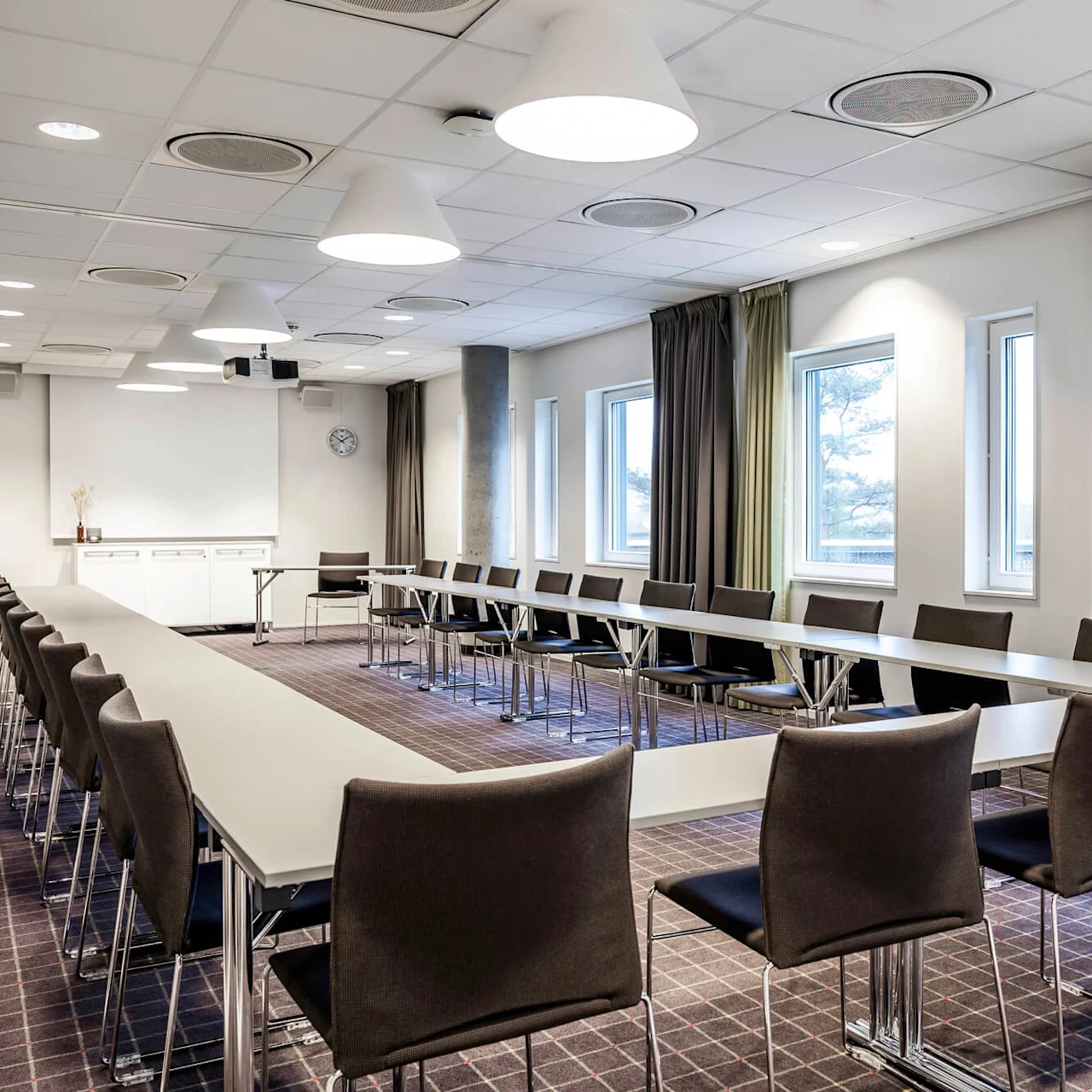 Et konferanserom med lange bord og stoler i en U-form, under lyse taklamper. Vinduer, en klokke og en projeksjonsskjerm er synlige.