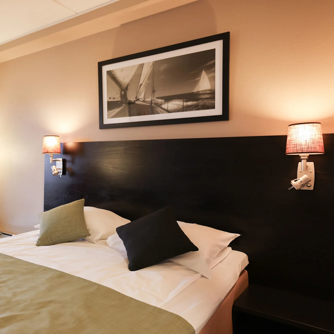 A neatly made bed with two pillows rests in a warmly lit hotel room with orange curtains and a framed sailing photograph above the headboard.