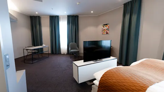 A modern hotel room with a bed covered by a brown quilt. A television faces the bed, and a desk with a chair is by the window with dark curtains. 