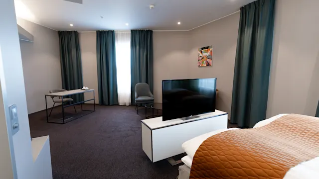 A modern hotel room with a bed covered by a brown quilt. A television faces the bed, and a desk with a chair is by the window with dark curtains. 
