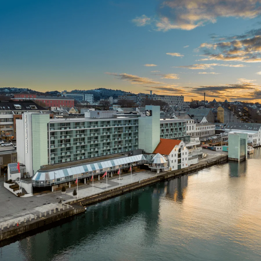 Ett hotell märkt "Quality Hotel Maritim" står bredvid en lugn strandlinje i solnedgången, omgivet av byggnader med en pittoresk stadsbild i bakgrunden.