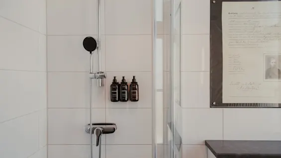 A modern shower with white tiles, a shower head, and three dark bottles of toiletries. A framed document hangs on the adjacent wall.