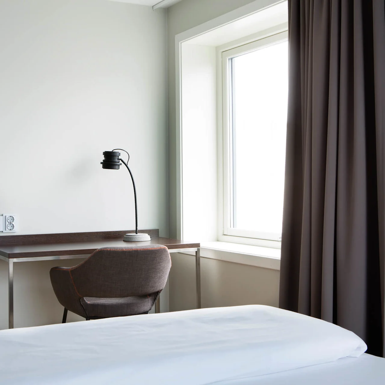 En stilren skrivebordslampe står på et trebord ved siden av en stol og et stort vindu med gardiner. Nøytrale farger, minimalistisk design.