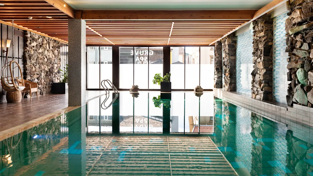 Indbydende indendørs poolområde på Quality Hotel Grand, Kongsberg, med naturligt lys, stenmure, hyggelig siddeplads og Grand Spa-reflektioner.