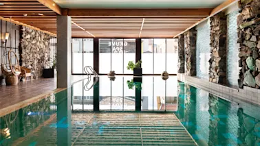 Innbydende innendørsbasseng på Quality Hotel Grand, Kongsberg, med naturlig lys, steinvegger, koselig sittegruppe og Grand Spa-refleksjoner.