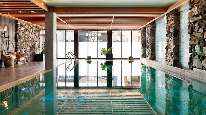 The inviting indoor pool at Quality Hotel Grand, Kongsberg, featuring natural light, stone walls, a cozy seating area, and Grand Spa reflections.