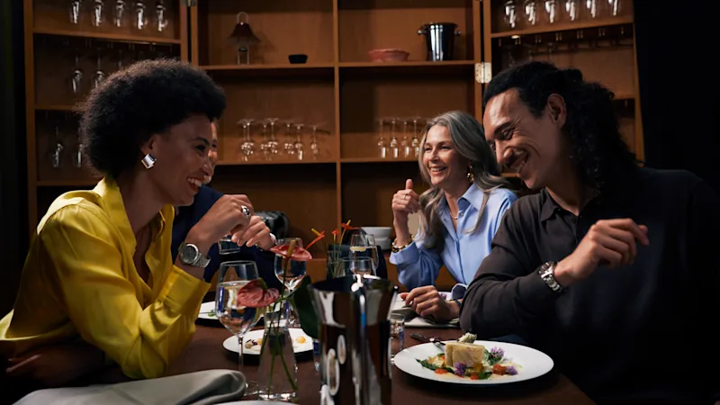 Kolme henkilöä istuu ruokapöydän ääressä vilkkaassa keskustelussa. Heidän ympärillään on lautasia ruokaa ja lasillisia juomaa, taustanaan hyllyt joissa on lasitavaraa ja pieni liitutaulu.