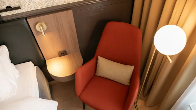 En rød stol med en beige pute står ved siden av en seng på et hotellrom. En rund gulvlampe lyser svakt, og et nattbord i tre har en veggmontert leselampe.