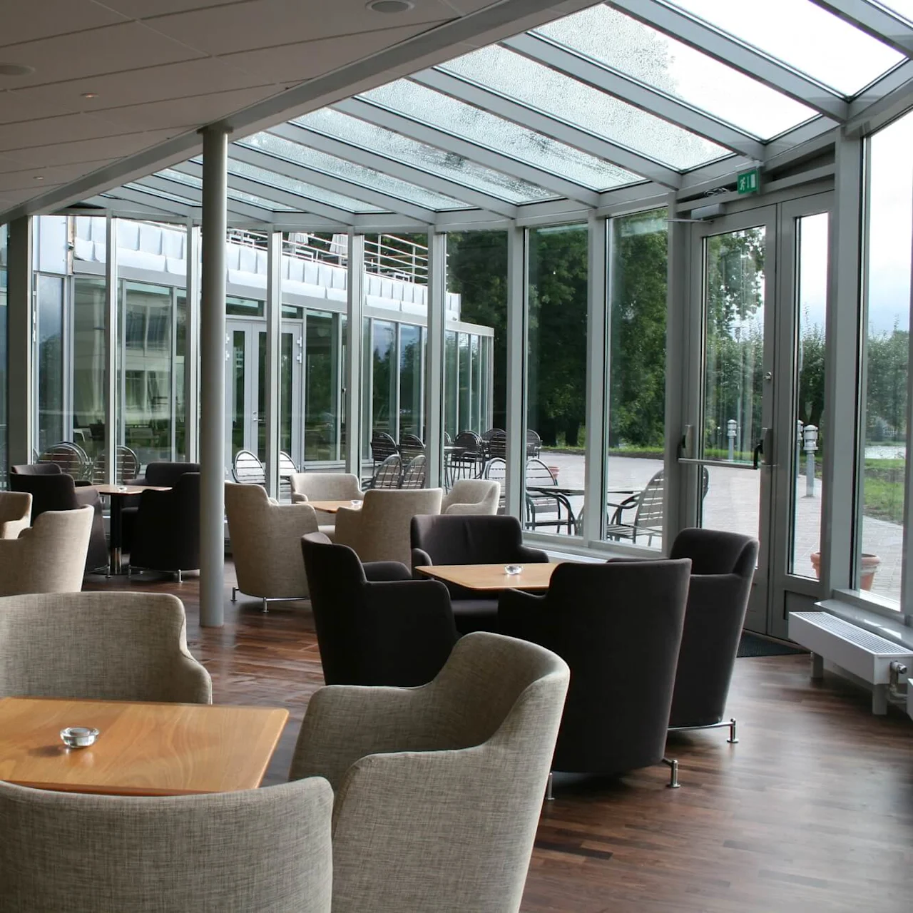 Borde og polstrede stole er arrangeret i et glasvæggområde. Sollys filtrerer gennem loftet og vinduerne, der har udsigt over en udendørs terrasse med flere siddepladser. Udgangsskilt synligt.