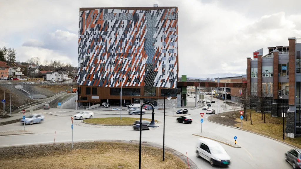 Moderne bygning med mønstret facade står ved en trafikeret vej med bevægelige biler, i en forstadsindstilling med skyet himmel.