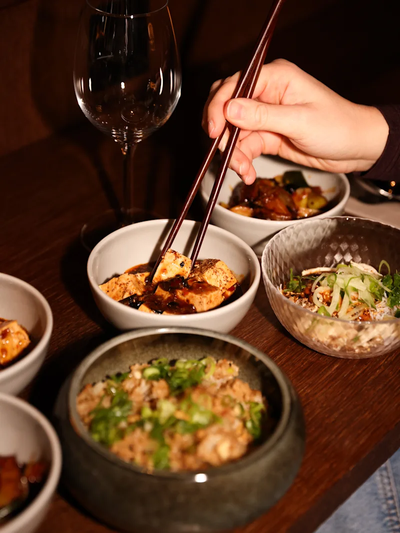 A hand uses chopsticks to pick up food from a bowl, surrounded by other small dishes and a wine glass on a wooden table.