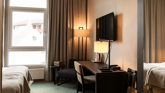 Et hyggeligt hotelværelse med skrivebord, TV og en komfortabel seng, hvor naturligt lys strømmer ind fra et stort vindue.