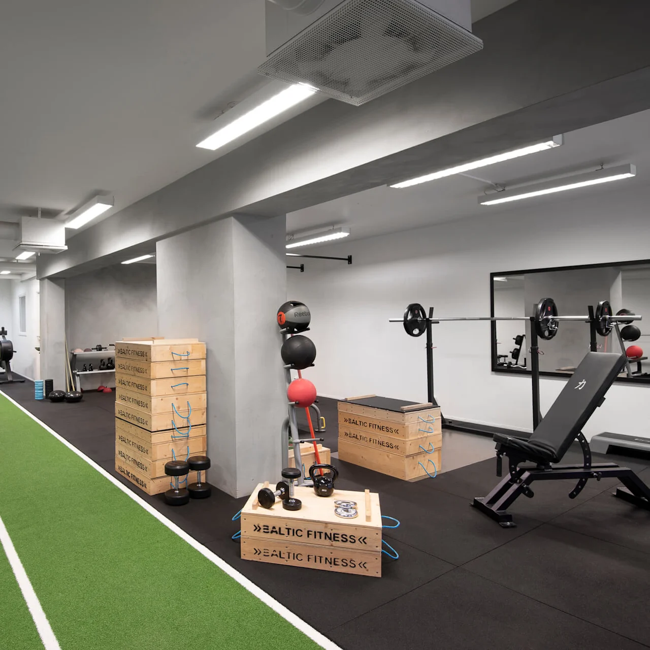 Gym med träningsutrustning; motionscykel, löpband och vikter är arrangerade runt en grön bana. Lådor märkta “BALTIC FITNESS” är staplade i närheten.