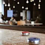 A shuffleboard with red and blue pucks on a sprinkled surface is in focus, set in a cozy lounge area with soft seating and overhead hanging lights.  