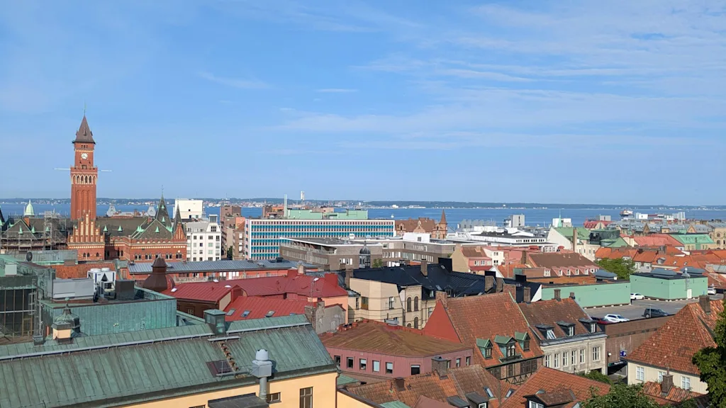 En klocktorn i röd tegelsten reser sig över stadens färgglada hustak, med blå himmel och avlägset hav i bakgrunden.