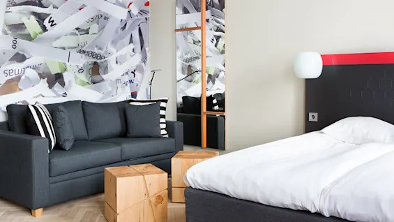 En grå sofa med svart og hvite puter står ved siden av en seng med hvitt sengetøy, i et rom med treterningbord. Abstrakt papirstrimler gir kunstnerisk sammenheng.