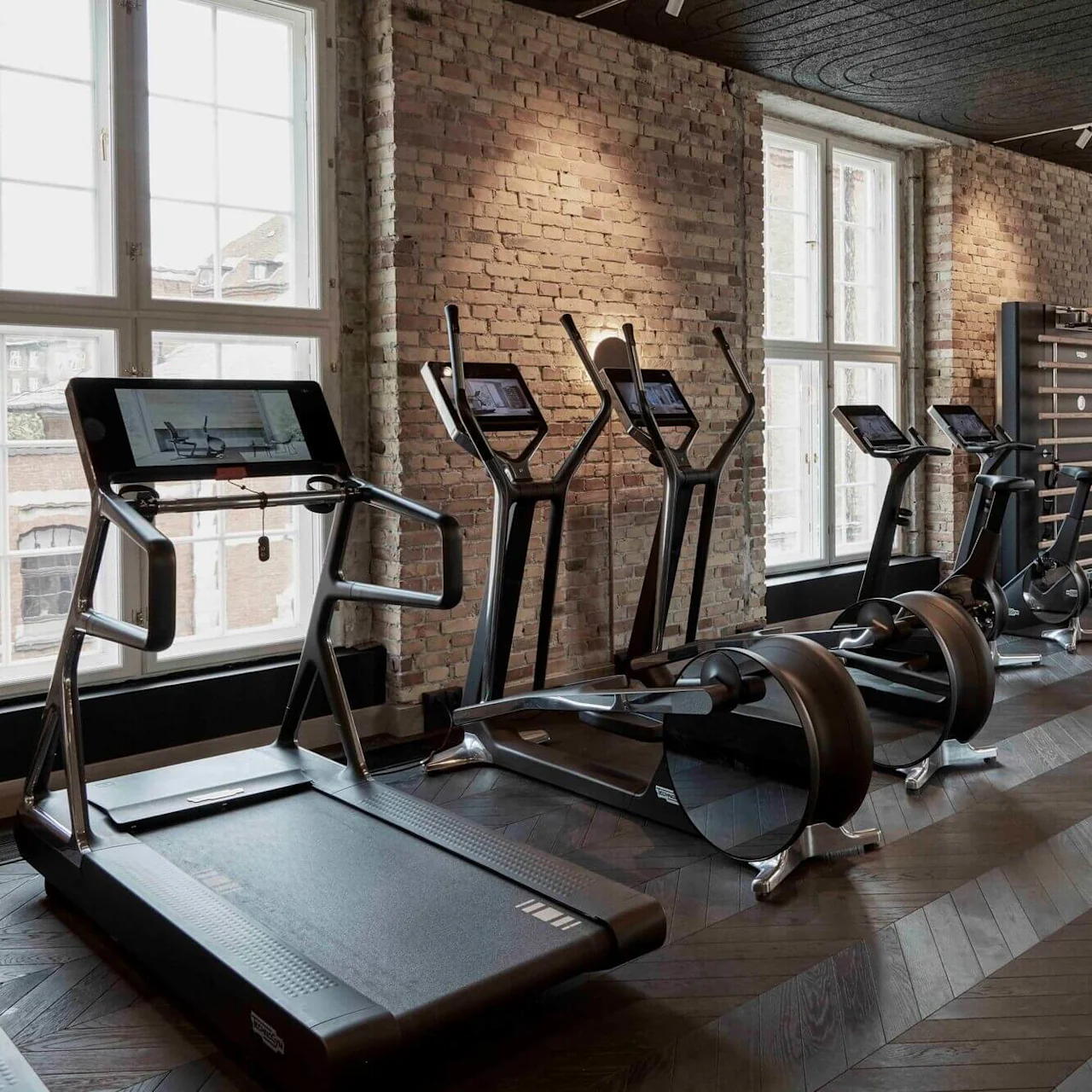 Treadmills and exercise bikes are neatly arranged in a modern gym with brick walls, wooden floors, and large windows providing natural light.