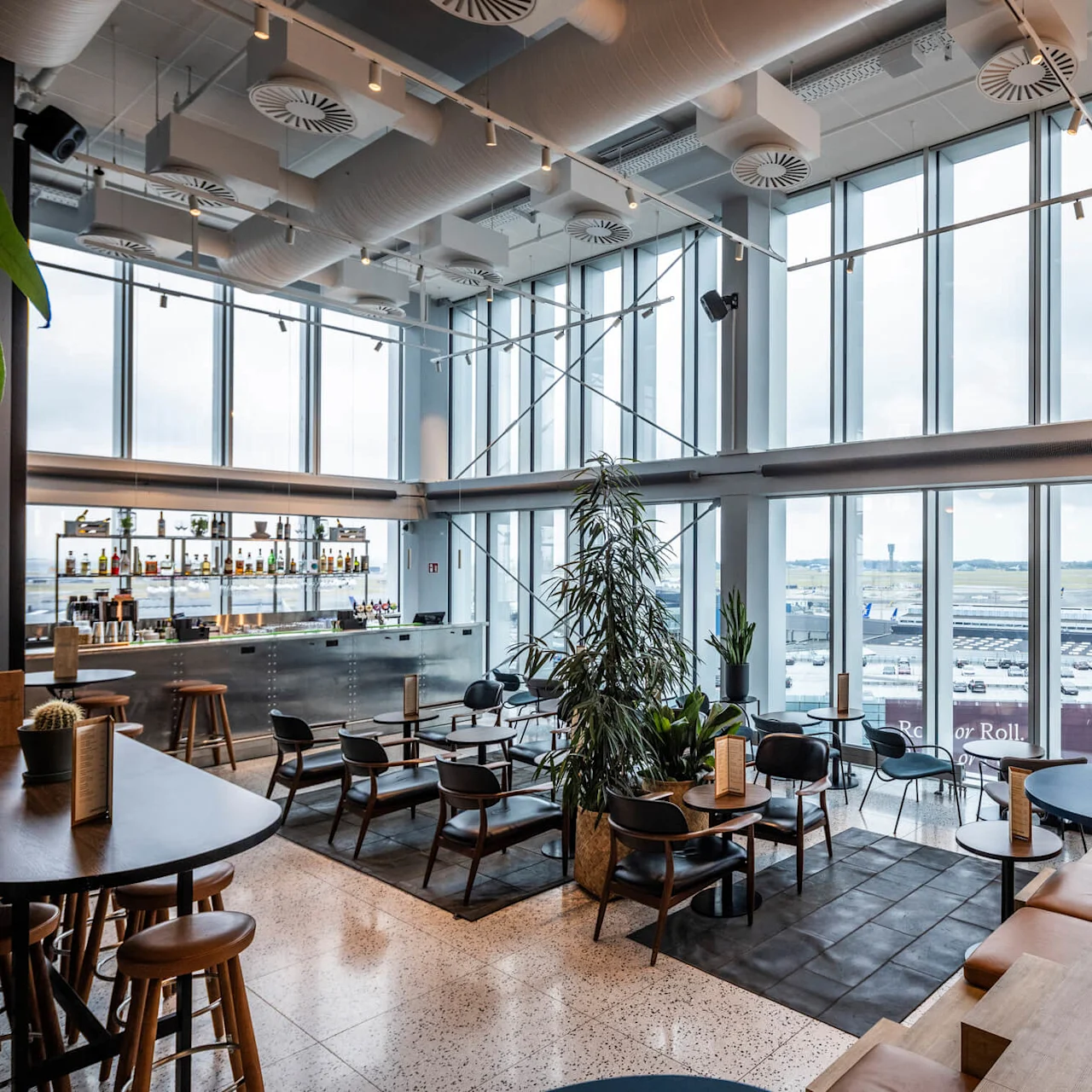 En romslig flyplasslounge med en bar, spredte sitteplasser og store vinduer med utsikt over parkerte fly og rullebanen. Området har moderne innredning og mye naturlig lys.
