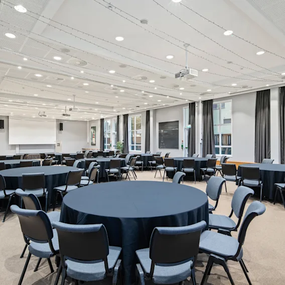 Runde bord dekket med mørke duker er omgitt av stoler i et romslig konferanserom med store vinduer og en projektorskjerm, som skaper en formell møteatmosfære.
