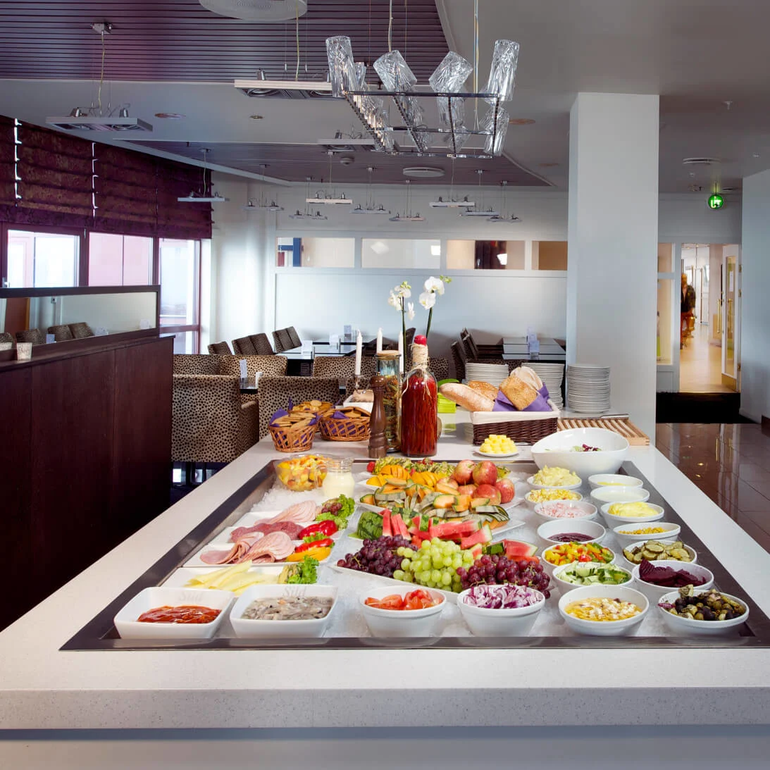 Et buffétbord er fyldt med forskellige retter som frugt, salater og pålæg. Det står i en moderne spisestue med dispensere og blomsterdekorationer.