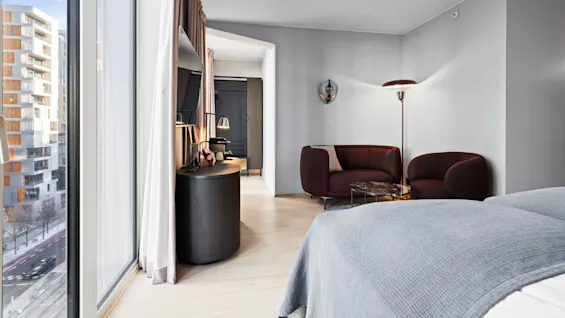 A modern bedroom with a grey bedspread, adjacent seating area with a maroon sofa, and large window overlooking an urban landscape with buildings. A TV and desk with lamps are present.