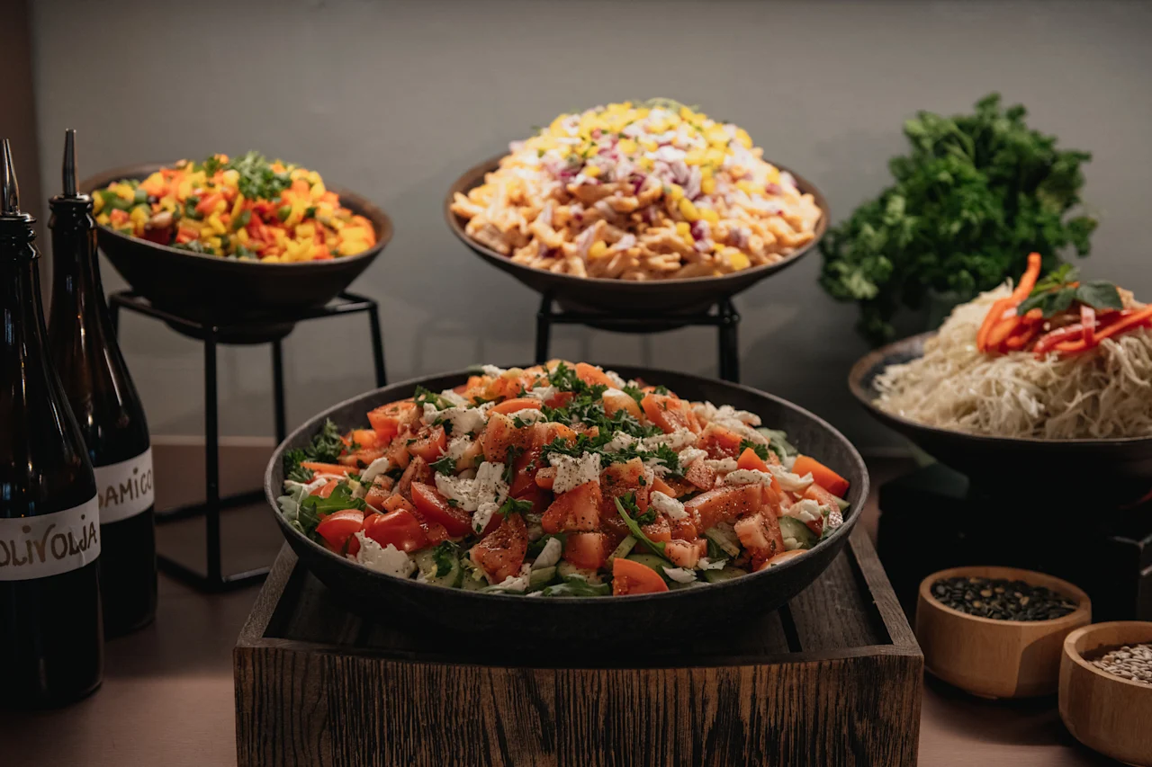 En livlig buffé med fräscha sallader: tomat och feta, pasta och nudlar. Flaskor med OLIVOLJA och frön syns också.