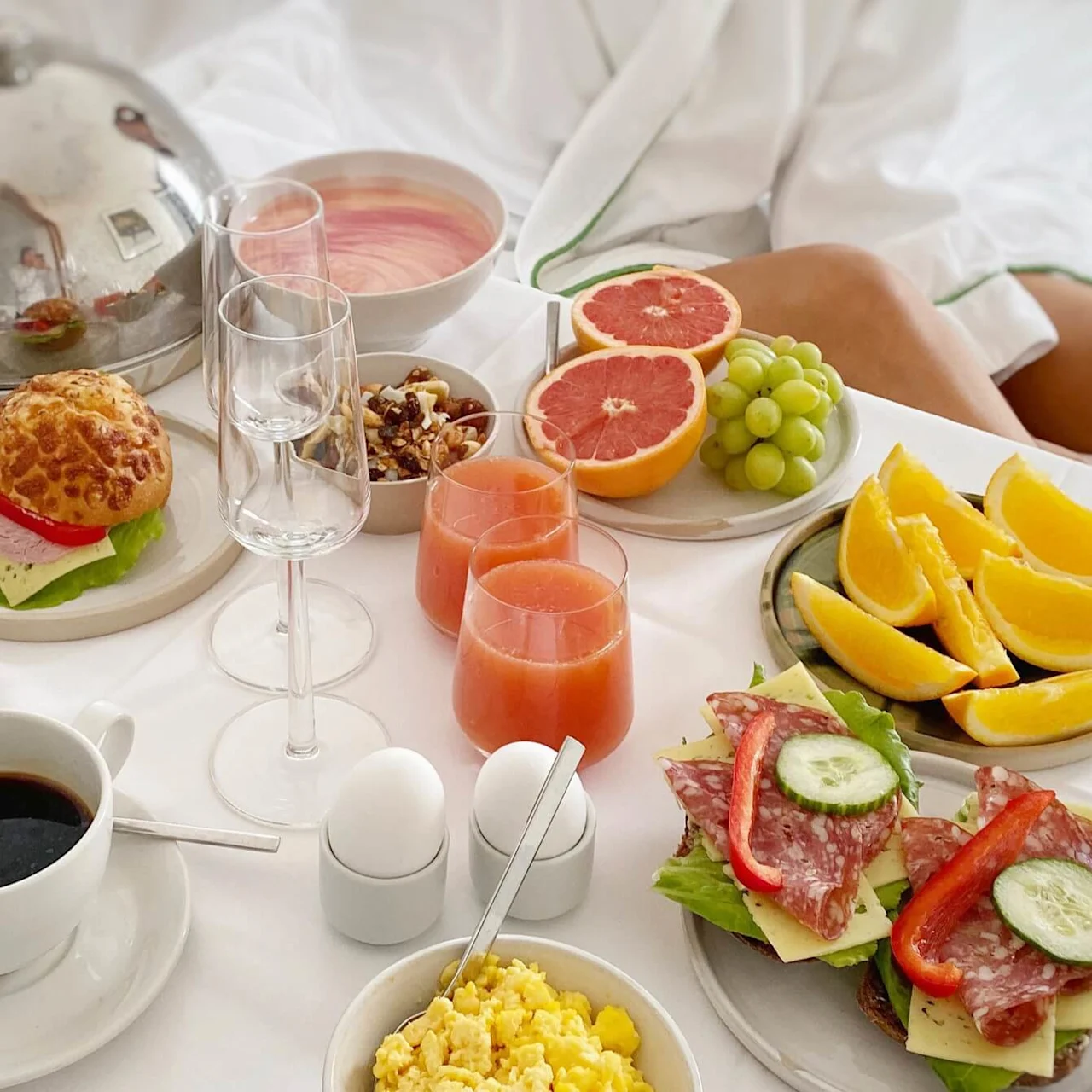 A luxurious breakfast spread features scrambled eggs, sandwiches, coffee, juice, and fresh fruit elegantly arranged on a white tablecloth, surrounded by two people in white robes.