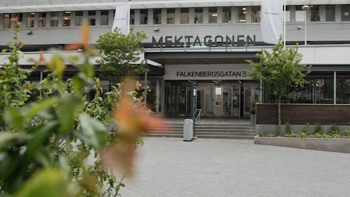 Bygningens indgang viser teksten "MEKTAGONEN" og "FALKENBERGSGATAN 3" over. Omgivet af træer, buske og brolagt gårdsplads. Hvide bannere hænger på facaden.