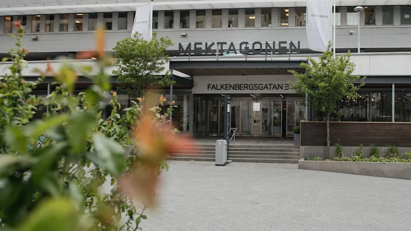 Building entrance displaying "MEKTAGONEN" and "FALKENBERGSGATAN 3" above. Surrounded by trees, bushes, and paved courtyard. White banners hang on the facade.