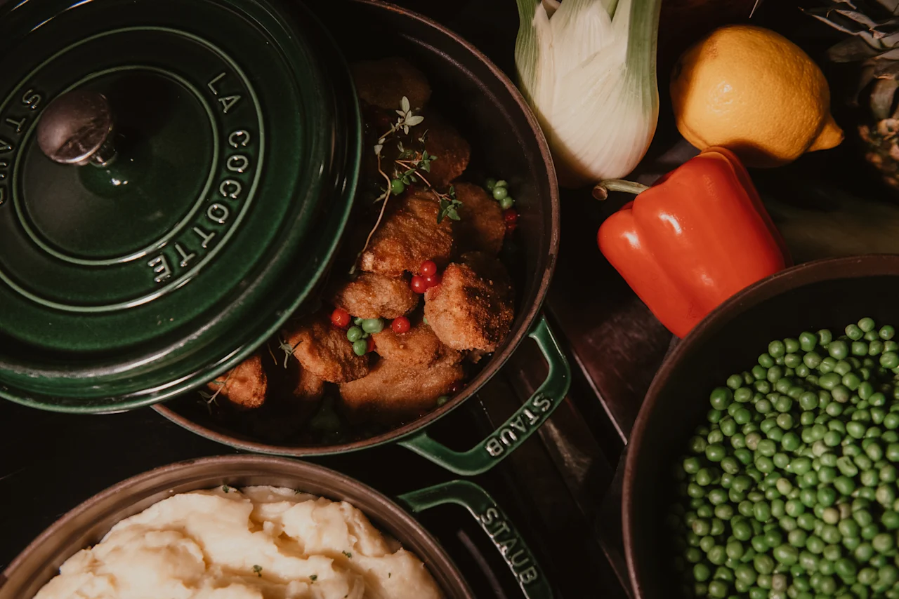 A delicious meal being served at Home Hotel Kung Oscar, featuring a pot of meat with LA COCOTTE, mashed potatoes, and peas. Fresh vegetables are on the side.